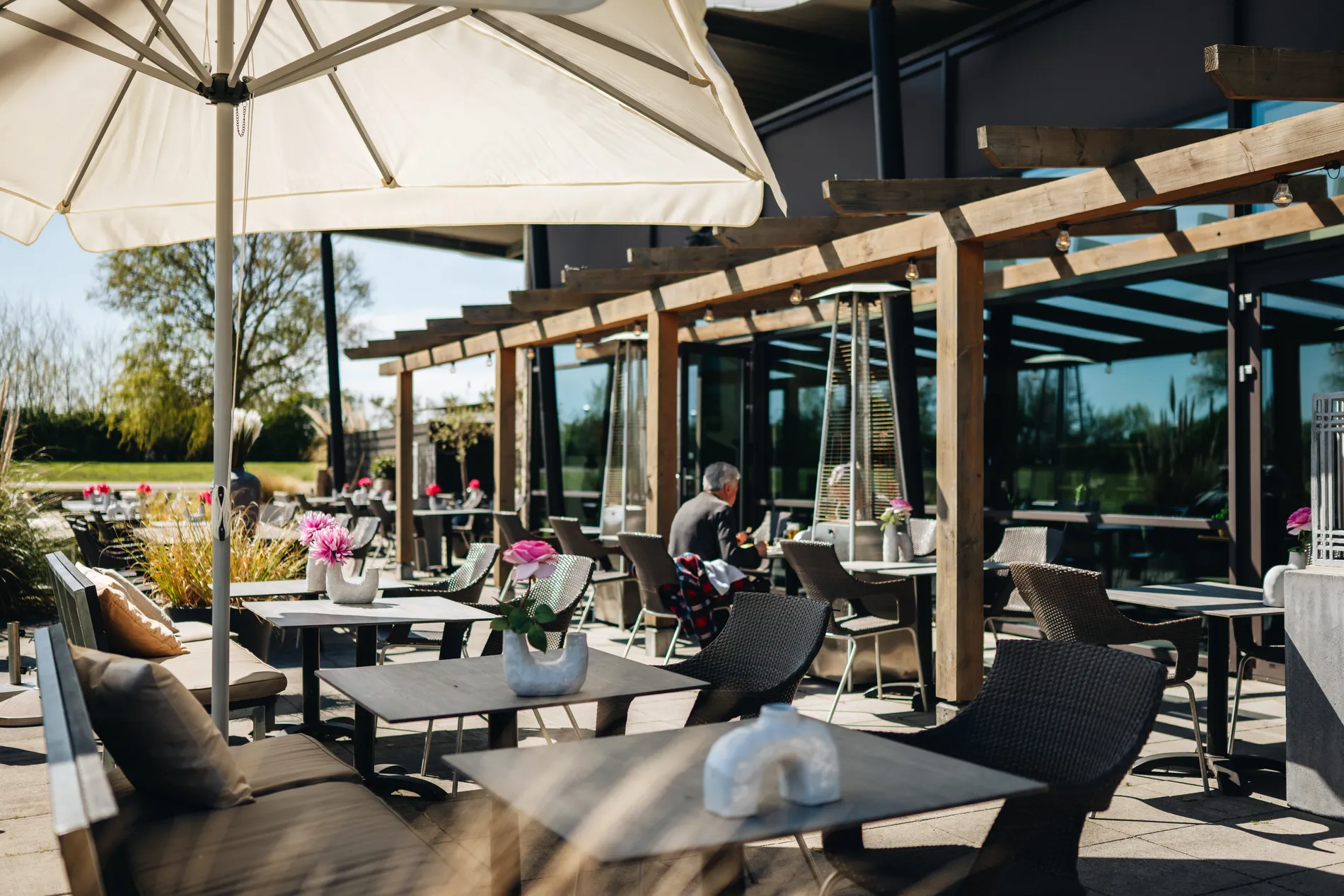 Een zonovergoten buitenterras met houten pergola's, terrasverwarmers en tafels gedecoreerd met roze bloemen in witte vazen.